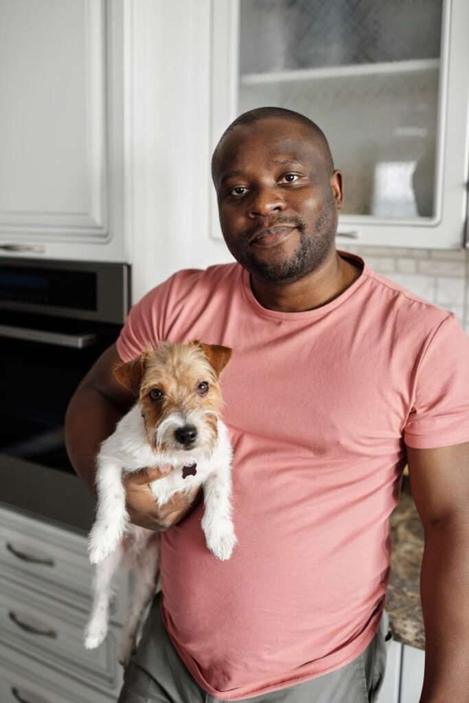 pexels-photo-4260745-4260745 A cheerful man in a pink shirt holds a dog in a modern kitchen, capturing a moment of togetherness.
